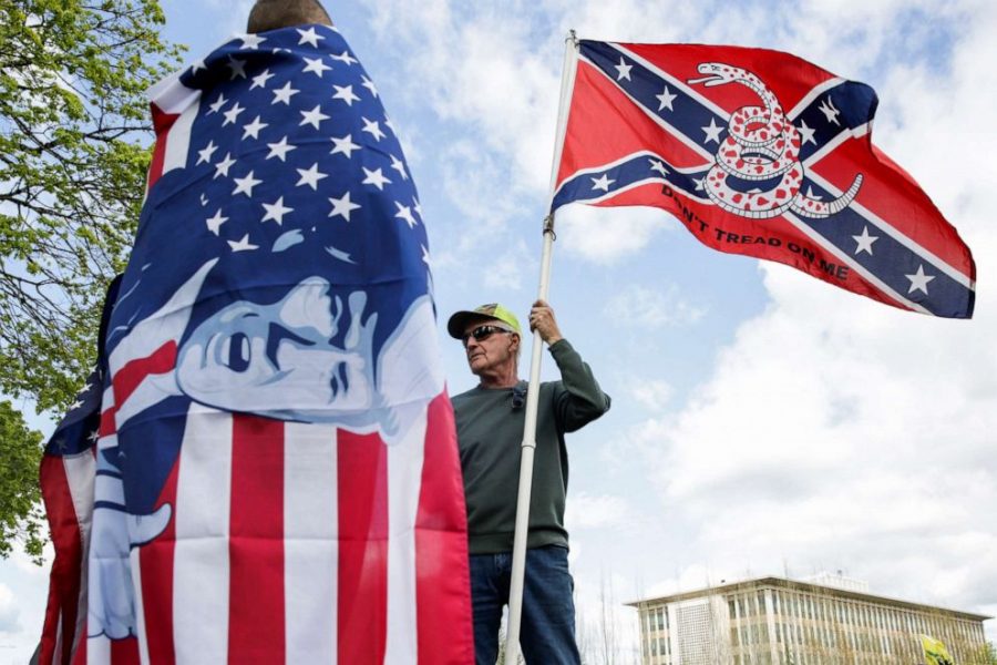 A right-wing protestor holds a flag that combines a Gadsden flag from the American Revolution with a Confederate flag from the Civil War. Though the two symbols, historically, contradict each other, they are now frequently seen together. He talks to another man holding an American Flag with President Trump’s face plastered over it.
