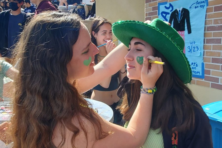 Sophomore Olivia Long paints sophomore Claire Kettwig's face with green paint, representing the St. Patrick's Day spirit. 