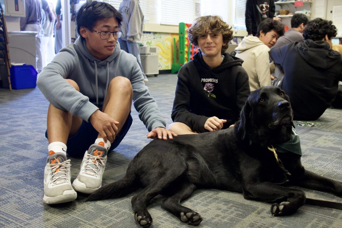 Two students sit with Meru, a black labrador. Meru started life as a puppy in the Guide Dogs for the Blind training program. He is now certified in “Pet Assisted Therapy” and is a “Canine Good Citizen.” 