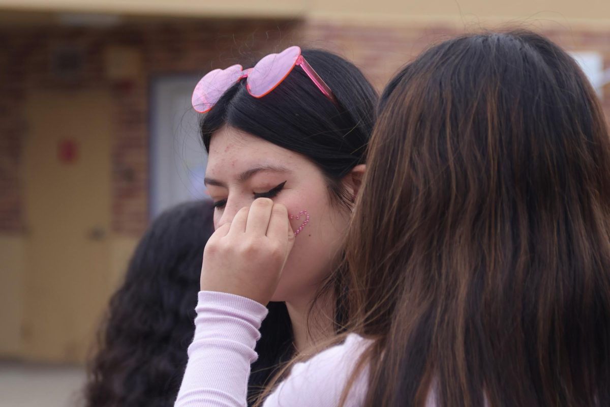 An ASB member applies gems to a students face at a booth. Many ASB members staffed stations in the quad to help with the many activities. At this booth, students picked designs representative of Valentines Day, such as a heart.