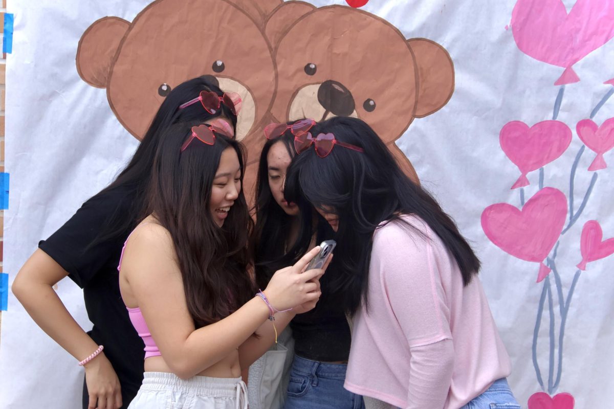 A group of girls huddle around a phone looking through photos taken with the hugs and kisses poster. Several groups came to the quad during lunch to participate in the various activities. Dressed in Valentines Day spirit, many stopped and posed with their friends to capture the moment.