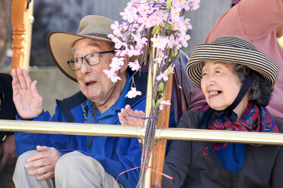 Two elderly people wave to the crowd. They sat in a trolley decorated with cherry blossoms. Similarly, those who didnt walk the parade waved from various floats, cars, or buses.