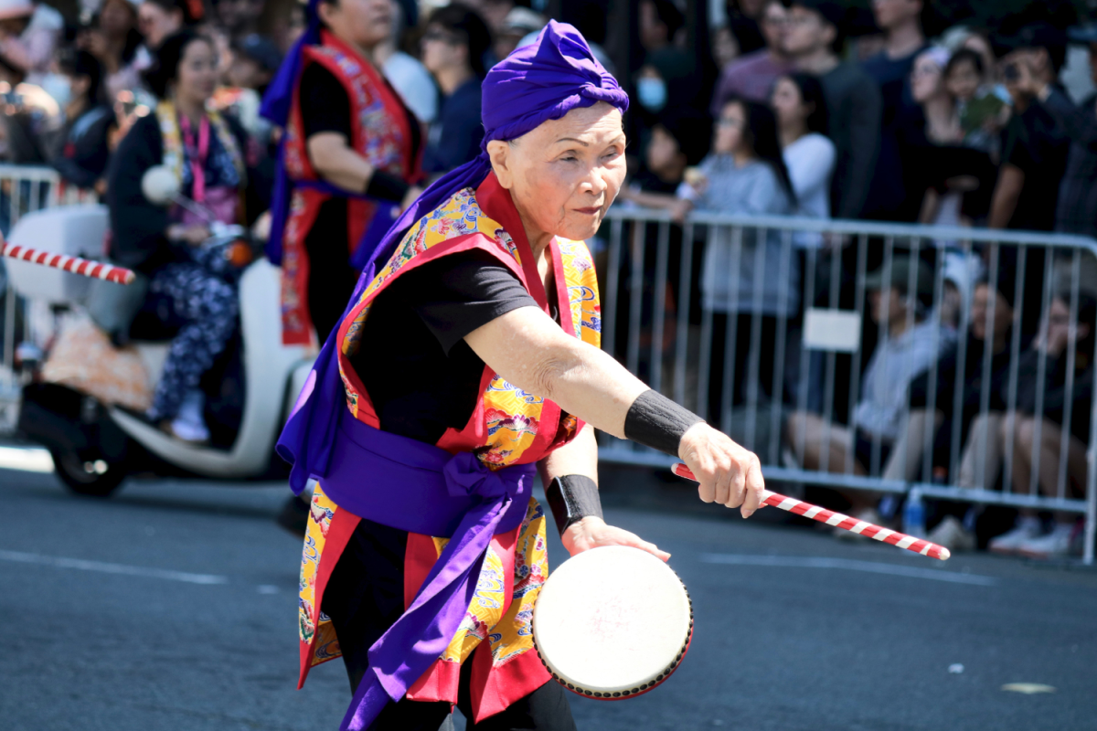 A woman performs a musical number with a handheld drum and wooden stick. Along with other members of her group, she meshed together fluid arm movements, footwork, and music. Her group walked down the hill and started their choreography once they were at the bottom.