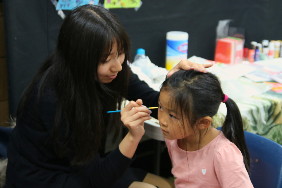 A volunteer does face paint on the side of a girls face. She used a mix of blue and white to create a floral design. There was a separate room in the bowling alley for face painting and crafts. 