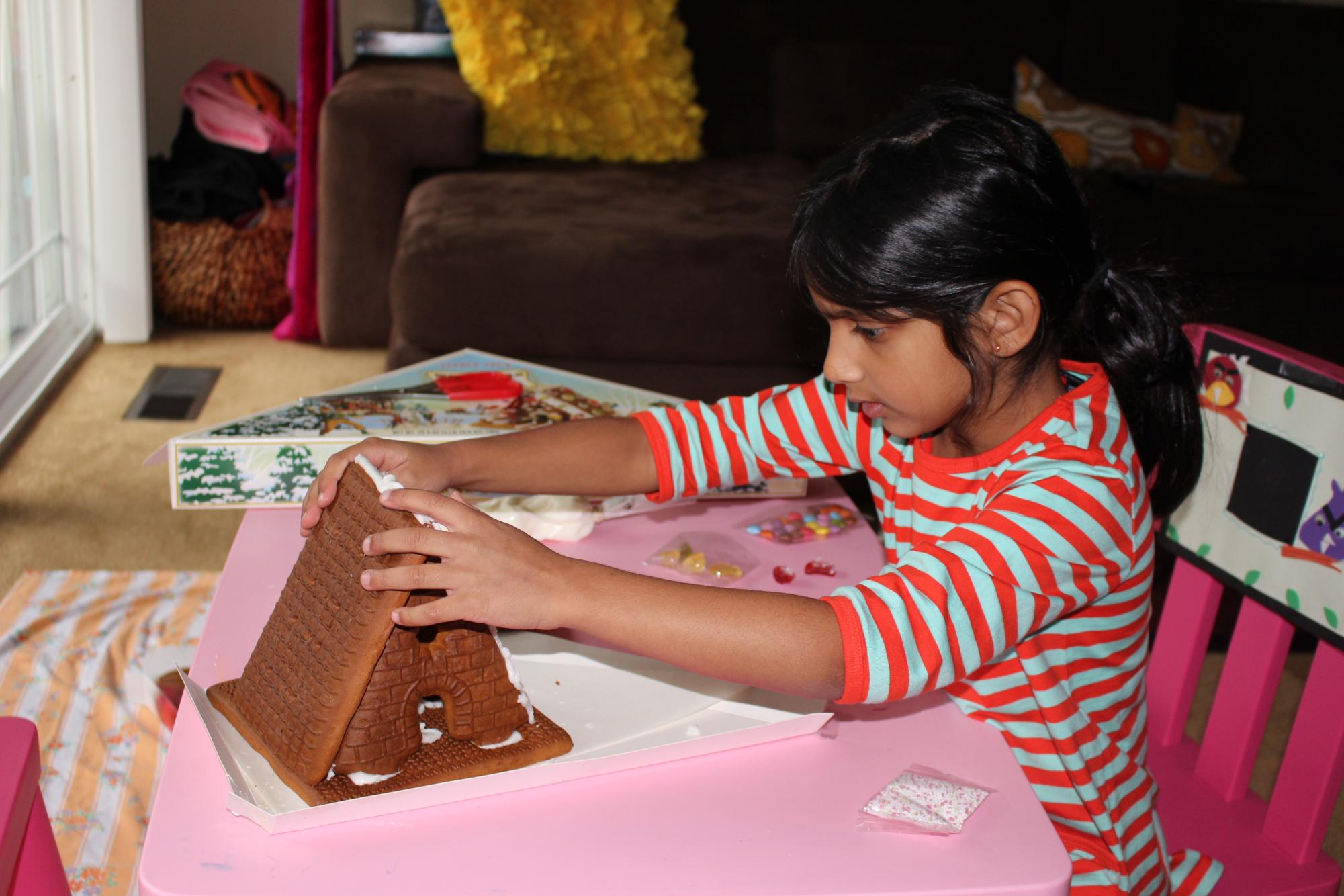 Anaya Goradia, at 3 years old, carefully builds her gingerbread house, focusing on making it just right. Her playful creativity shines as she decorates at her pink table, surrounded by candy.