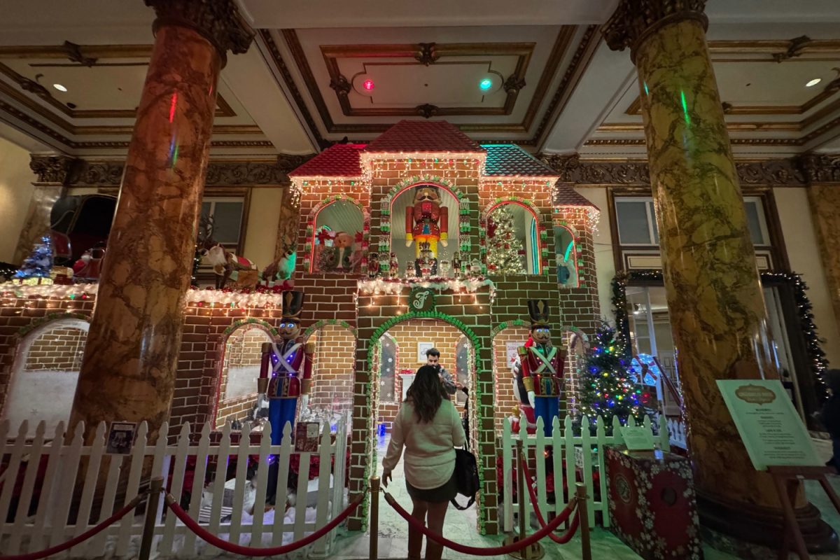 A woman stands in front Fairmonts life sized gingerbread house admiring it as she poses for a photo with a Christmas tree behind her. The house took 520 hours to build using 8,000 gingerbread bricks and over 2,000 pounds of candy.