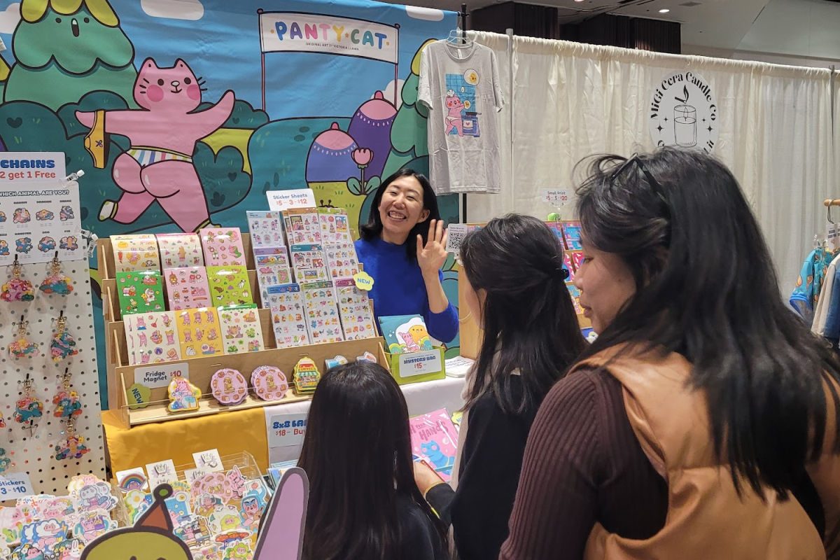 Victoria Llama, an illustrator and the owner of Panty Cats, stands in her booth. The booth displays all her cartoons in the form of pins, t-shirts, stickers, and much more.