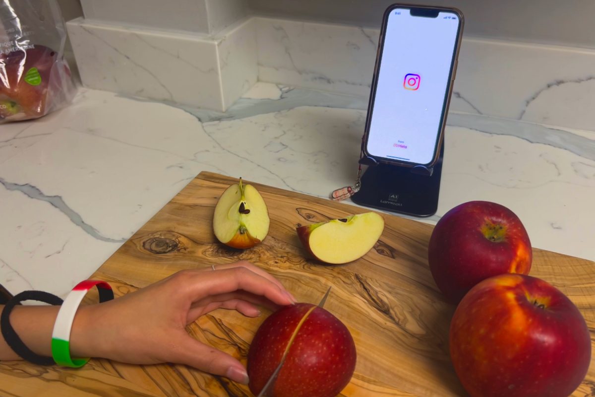 A person cuts apples for a meal while looking at a phone displaying the Instagram logo, illustrating how many people today use social media to learn life skills such as cooking and baking. "The pros of using videos are that you can see what your baked good is supposed to look like as you're making it," said Victoria Black, a Carlmont freshman. 