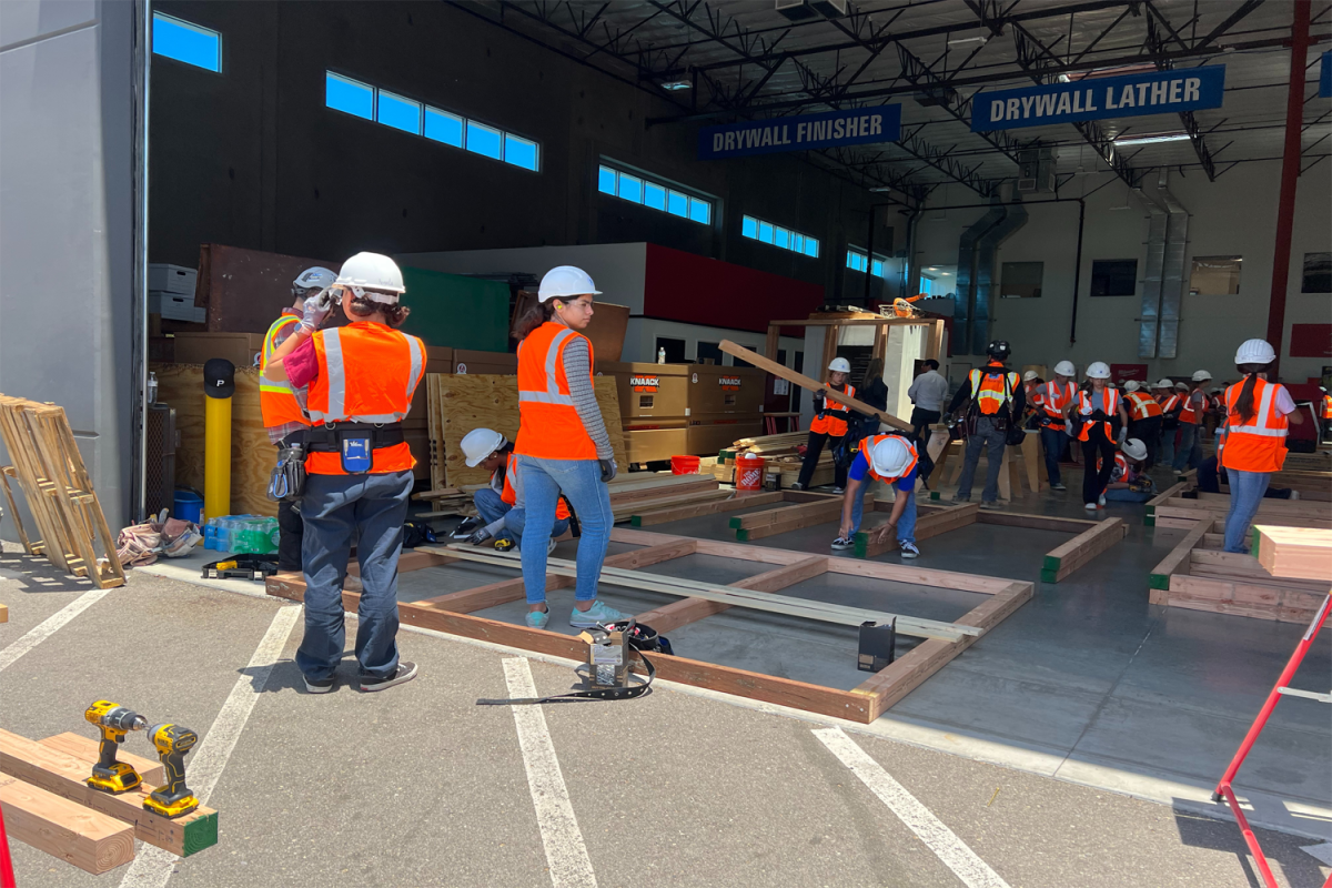 A group of women labor on the construction field. They haul wood and set it down on the floor to create a frame. Despite construction seeming like an excruciating task that only the strongest can handle, typically those who are male, women of all sizes are capable of handling tools and procedures during a build.
