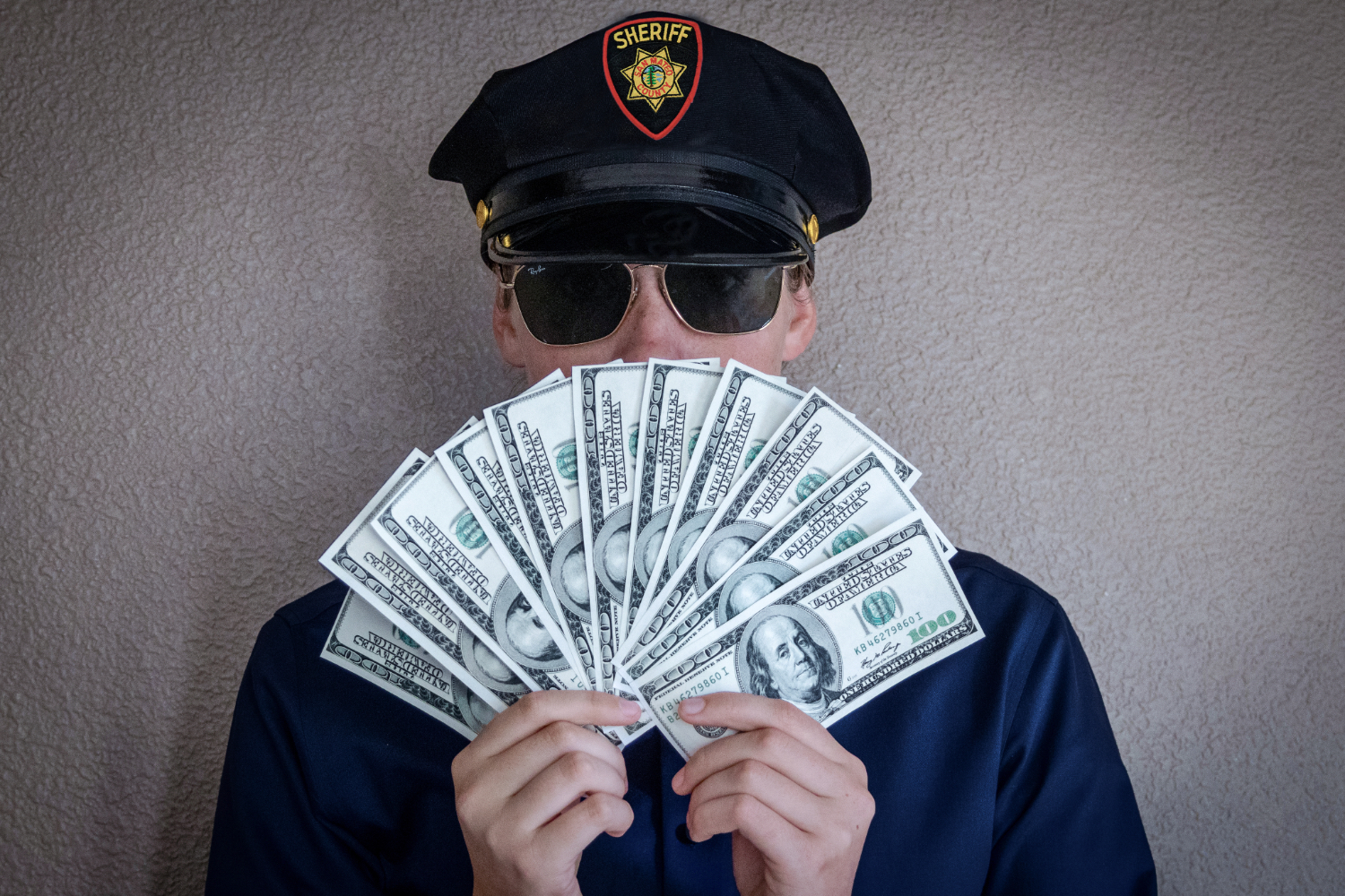 A law enforcement officer fans out money in front of his face, concealing his identity. For nearly two decades, the San Mateo County Sheriff's Department has been plagued with sheriffs corrupted by money and power. 