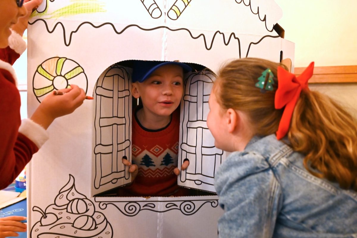 A boy talks to the children through the open window of a cardboard gingerbread house. The children were given crayons and markers to decorate the gingerbread house together and play inside it, one activity that encouraged creativity and new friendships at Belmont’s Winter Wonderland.