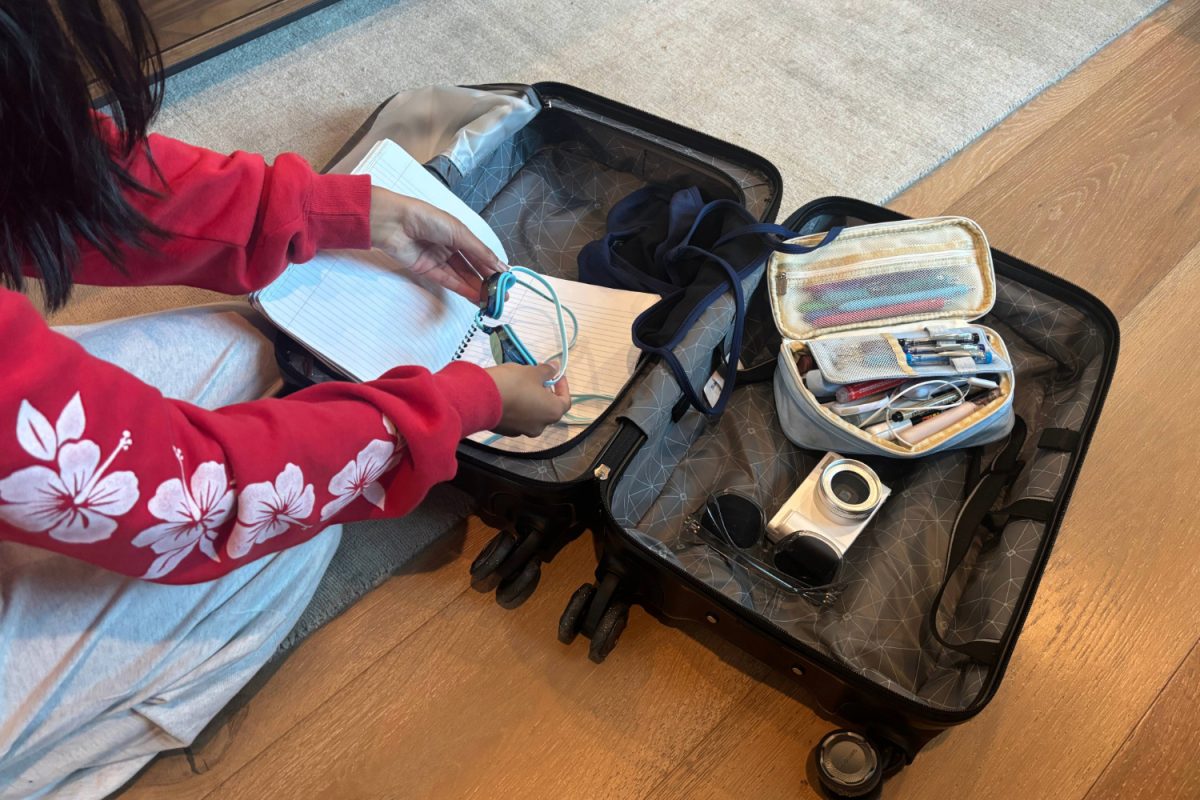 A student packs their suitcase full of items like a camera, sunglasses, and swim goggles. The variety of items represents the different ways students choose to spend their time over break, whether it's on vacation, studying, playing sports, or more.