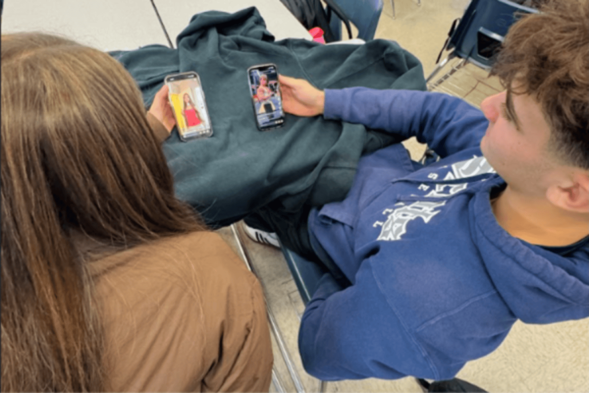 Two students sit at the same table looking at their phones. Each students sees a different message about how they should look on the same social media platform.